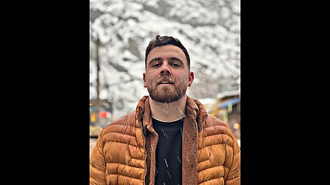 Potrait of the 26-year-old protester Erfan Soltani stands outdoors in snowfall, wearing a brown puffer jacket. Snow-covered mountains blur in the background.
