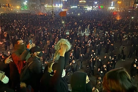 Crowd of masked protesters, mostly in dark clothing, gather at night on a street. Raised fists and signs convey determination and solidarity.