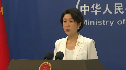 China's spokesperson Mao in a white suit speaks at a podium with two microphones. The blue backdrop displays Chinese characters and "Ministry of". A red flag is partially visible.