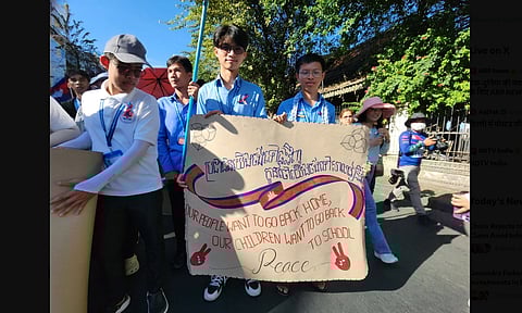 A group of people hold a sign reading "Our People Want to Go Back Home" at a peaceful protest. The mood is hopeful, with sunny weather and trees behind.