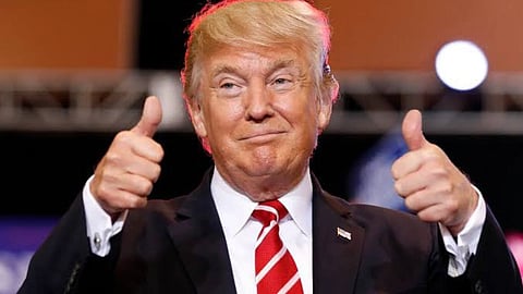 US President Donald Trump in a suit with a red and white striped tie gives a double thumbs-up. The background is blurred, and he has a confident expression.