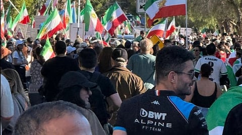 A large crowd gathers outdoors, carrying numerous Iranian flags. Trees line the background under a clear sky, creating a spirited and peaceful protest atmosphere.