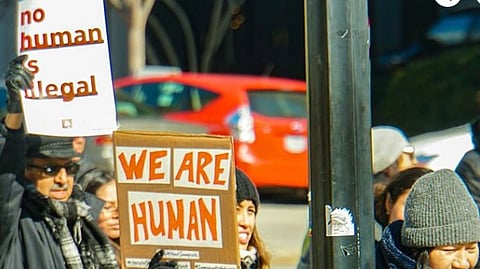A diverse group of protestors holds signs saying "No human is illegal" and "We are human" during a sunny outdoor rally, conveying unity and determination.