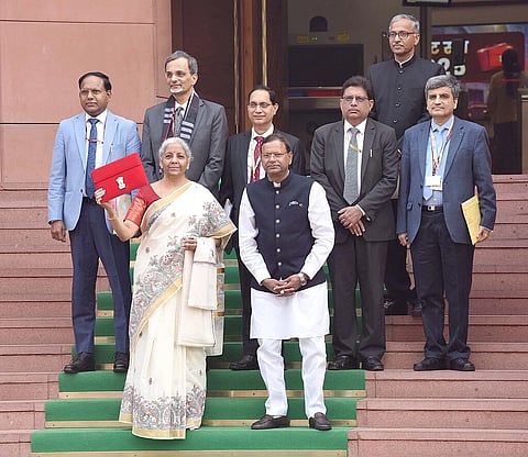 FM Sitharaman stands with other members of the Finance Ministry on carpeted stairs holding a red envelope.