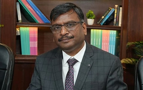 A man in a gray plaid suit and glasses sits in an office, with colorful books and a small plant on the wooden bookshelf behind, conveying professionalism.