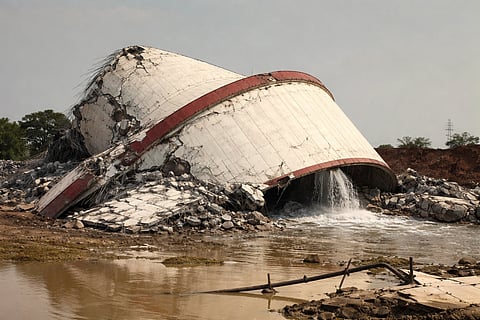 Collapsed water tower with a broken concrete structure and red bands, releasing water into a muddy pool. Overcast sky, somber and dramatic scene.