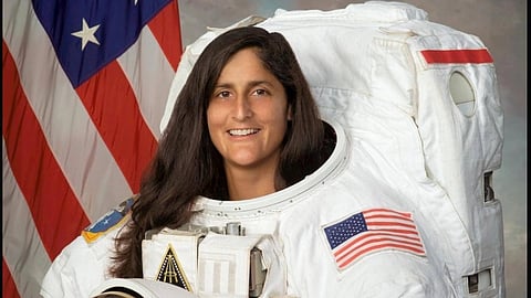 NASA astronaut Sunita Williams in a spacesuit holding a helmet under one arm, smiling. The American flag is in the background, conveying a sense of pride and achievement.