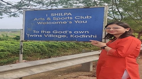 A woman in a red-orange kurta stands smiling next to a blue sign. The sign welcomes visitors to "God's own Twins Village, Kodinhi." Lush greenery surrounds them.