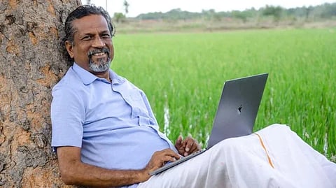 Sridhar Vembu in a light blue shirt and white dhoti leans against a tree, smiling while using a laptop in a lush green field, conveying a peaceful, relaxed mood.