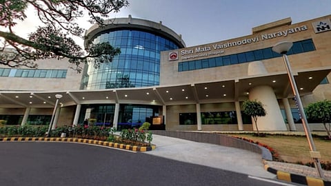 Modern hospital building with a circular glass entrance and beige stone facade. The sign reads "Shri Mata Vaishno Devi Narayana Super specialty Hospital." The foreground features a landscaped garden and a driveway.