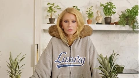 Hollywood actress Gwyneth Paltrow in a cozy, fur-collared "Luxury" hoodie stands in a kitchen. Surrounding her are pineapples, a watermelon, cucumbers, and a lime on a marble countertop. She appears calm and focused, with potted plants on a shelf in the background, adding a fresh, organic