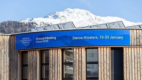 A wooden building with a banner reads "World Economic Forum, Annual Meeting Davos 2026, Davos-Klosters, 19-23 January." Snowy mountains in the background.