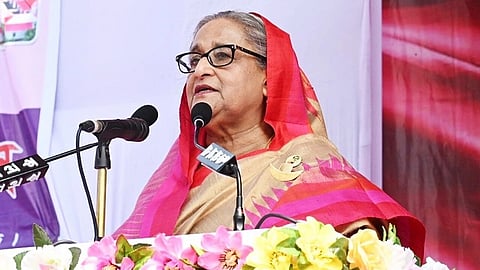 Bangladesh’s ousted former Prime Minister, Sheikh Hasina in a gold and pink sari speaks confidently at a podium adorned with colorful flowers. The background features abstract colors and patterns.