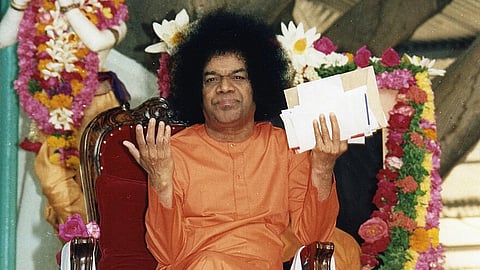 Sathya Sai Baba at Brindavan, sitting on a chair covered with garlands, wearing an orange kurta, holding his hands up while holding several letters.