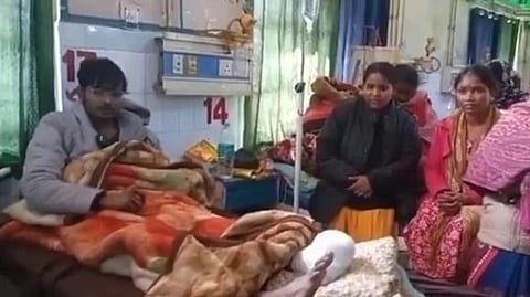 A man lies in a hospital bed covered with warm blankets, surrounded by three concerned women. The room has green curtains and medical equipment