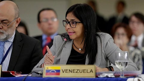Venezuelan interim President Delcy Rodríguez in glasses speaks into a microphone at a conference, sitting beside a sign labeled "Venezuela." Others are blurred in the background.