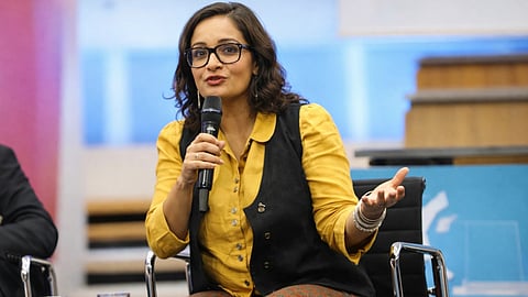 A woman in glasses and a yellow shirt with a black vest speaks into a microphone. She gestures expressively, seated on a stage, conveying engagement.