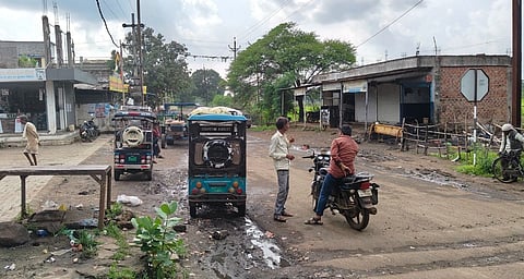 a village locality, a kaccha road, an e-rickshaw parked on the left side of the road, and a bike adjacent to it, mounted by the rider, the rider is talking a person to his left, probably the auto driver, houses/shops at both sides of the road, trees in the background, poles and wires at the left roadsie