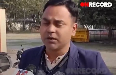 A man in a blue blazer and white shirt is speaking into a microphone, with a serious expression. The setting is outdoors with trees and a parked bicycle. The word "ON RECORD" is visible at the top.