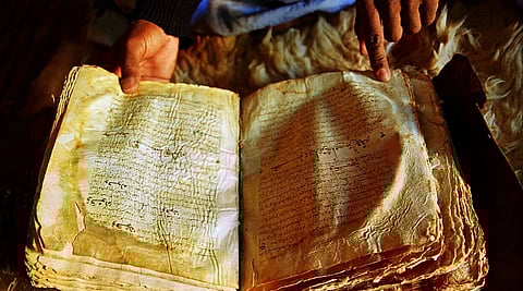 Image of nine-century-old Quran bound in gazelle skin in the library of Chinguetti, Mauritania, Africa.
A person's hands gently hold and point to an ancient, worn manuscript with aged, yellowed pages covered in faded, intricate script, evoking a sense of history.