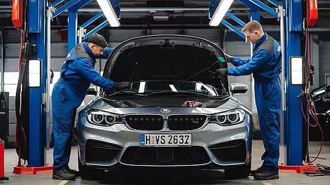 Two mechanics in blue uniforms work on a silver car with its hood open in a well-lit garage, conveying focus and professionalism.