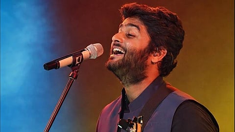 Singer Arijit Singh joyfully sings and plays guitar on stage, illuminated by vibrant blue and orange lights, conveying a lively and energetic performance vibe.