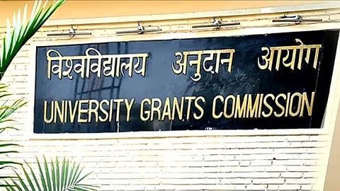 In the image a black banner is shown where University Grants Commission is written in Hindi and English both and there is a tree alongside that