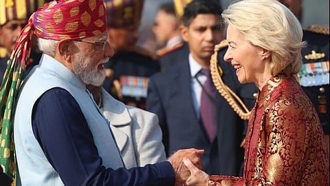 PM Narendra Modi in a colorful turban and European Commission President Ursula von der Leyen in a red patterned coat shake hands, smiling warmly. Uniformed guards with decorative hats stand in the background.