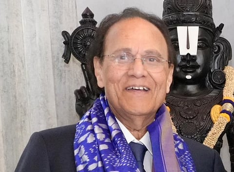 Image of radiation oncologist Dr. Nori Dattatreyudu with scarf smiling, standing in front of a detailed stone deity statue adorned with garlands.