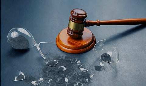 A judge's gavel rests beside a broken hourglass on a dark surface, with scattered glass pieces. The image conveys urgency and finality.