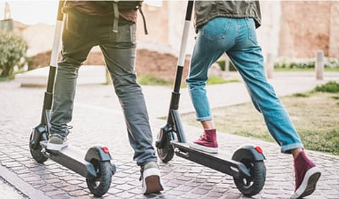 Two people ride electric scooters on a cobblestone path. They're casually dressed in jeans and sneakers, suggesting a leisurely, urban setting.