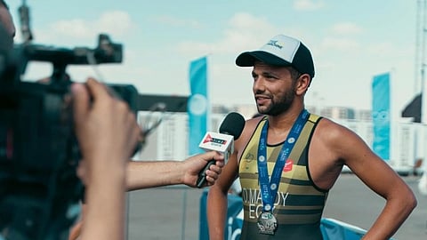 A man in a sports attire and cap is being interviewed after finishing a race. He wears a medal and smiles, exuding a sense of achievement.