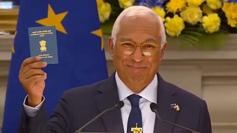 European Council President Antonio Costa in a suit, holds up his OCI card. Behind him, a European flag and yellow and blue flowers are visible, conveying a celebratory tone.