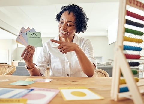 A smiling woman in a white shirt sits at a table, holding and pointing at colorful vocabulary cards. An abacus is visible on the right, suggesting a teaching setting.