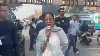 Mamata Banerjee is shown adrressing the people, she is wearing white saree and many man are going from behind her