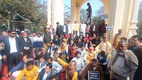 A group of people sit and stand at a public structure, holding signs in an organized protest. The scene is lively with a statue in the background.