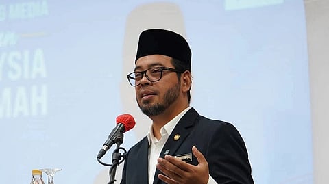 Dr Zulkifli Hasan in formal attire and a black cap speaks at a podium with a microphone, set against a backdrop of conference slides. He appears focused and engaged.