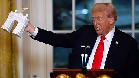 Donald Trump in a suit and red tie holds up a white model of a classical arch monument. He stands at a podium in a room with elegant decor. The mood is formal.