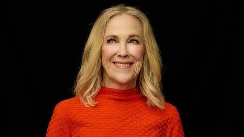 Catherine O'Hara with blonde hair in a bright red textured turtleneck and dark leather skirt stands against a black background, exuding warmth and confidence.