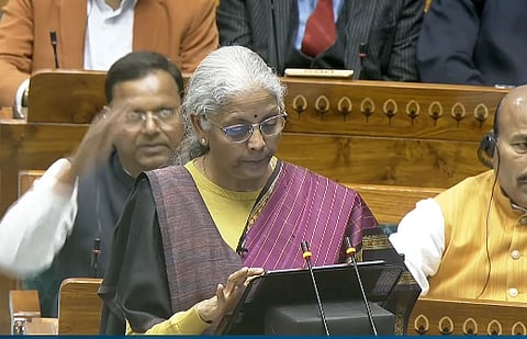Nirmala Sitaraman with gray hair and glasses speaks at a podium in an official setting. She wears a purple and yellow sari. Seated people are blurred behind he, presenting budget.
