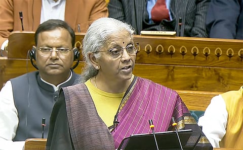 A woman in a purple sari speaks at a podium in a formal setting, surrounded by seated individuals in business attire. She appears focused and engaged.