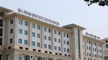 Large, modern building with cream facade and multiple windows. Sign reads "All India Institute of Ayurveda." Clear sky, calm and institutional atmosphere.