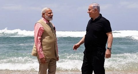 Image of PM Modi and Israel PM Benjamin Netanyahu are standing on a beach, facing each other and smiling. One is in a beige suit and the other in a black outfit. Waves crash in the background.