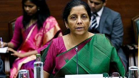 Image of FM Nirmala Sitharaman in a green and pink sari speaks at a conference table, looking attentive. Two people take notes behind her,