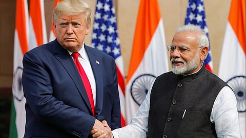 Indian PM Narendra Modi and US President Donald Trump in formal attire shake hands, standing in front of a backdrop of alternating Indian and American flags. The mood is formal and diplomatic.