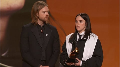 Billie Eilish and Finneas stand on stage at an awards ceremony. Billie Eilish holds a Grammy, speaking into a microphone, while both wear formal black attire.