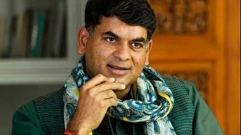 Journalist Saurabh Dwivedi in a green shirt and blue vest, smiling warmly, wearing a colorful peacock-patterned scarf indoors. The setting appears relaxed and thoughtful.