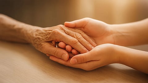 Two hands gently clasp an elderly hand in a warm, comforting embrace on a wooden table. The scene conveys empathy, care, and connection.