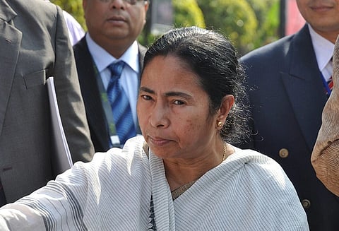 Image of CM Mamata Banerjee in a white saree with black stripes standing outdoor among a group of men in suits. She has a serious expression, and greenery is visible behind her.