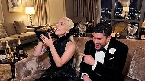 Lady gaga in a black feather dress kisses a Grammy award, while Bad Bunny in a tuxedo laughs and points at her. The room is elegantly decorated.